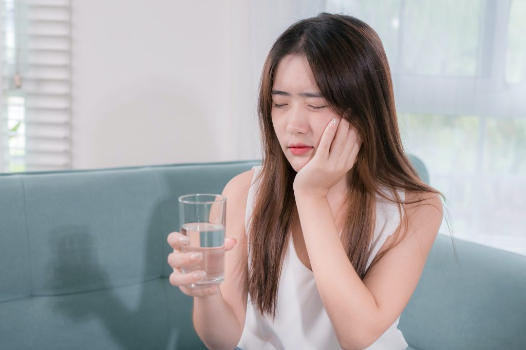 Asian woman has sensitive teeth after drinking cold water on sofa in living room at home. concept de